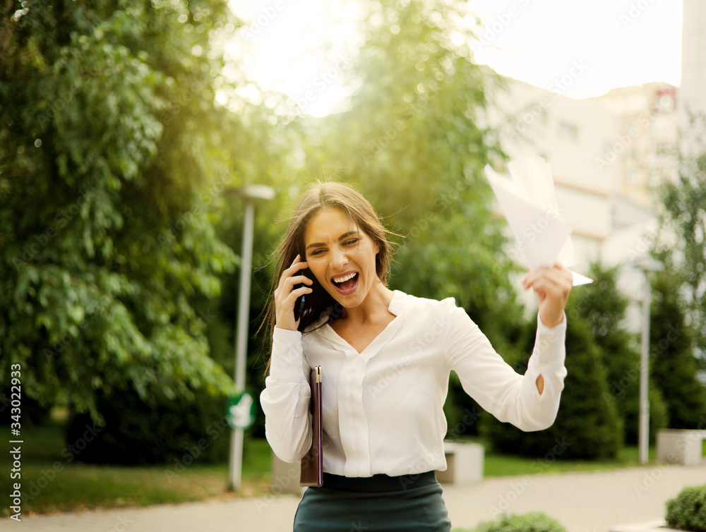 Brought to hysteria, angry business woman dressed in a white blouse and a skirt screams in the phone while holding sheets of paper outdoors in the city Brought to hysteria, angry business woman dressed in a white blouse and a skirt screams in the phone while holding sheets of paper outdoors in the city