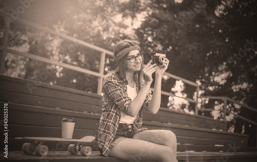 Funny female skater dressed in stylish clothes sits in the stands in skatepark while holds retro camera and listens to music in earphones at bright sunny day