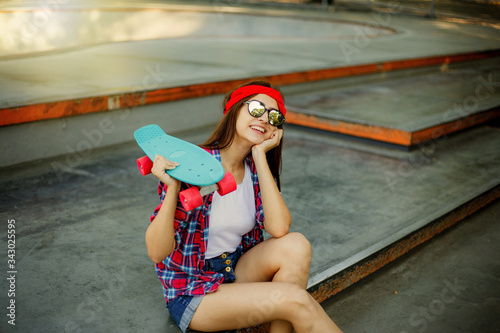 Portrait of stylish skater woman in skatepark. Attractive girl in red plaid shirt, mirrored sunglasses, shorts rests with a skate