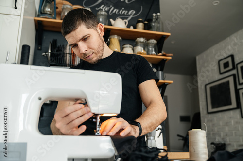 Attractive seamstress man or clothing designer works with a sewing machine and black cloth. Remote work.