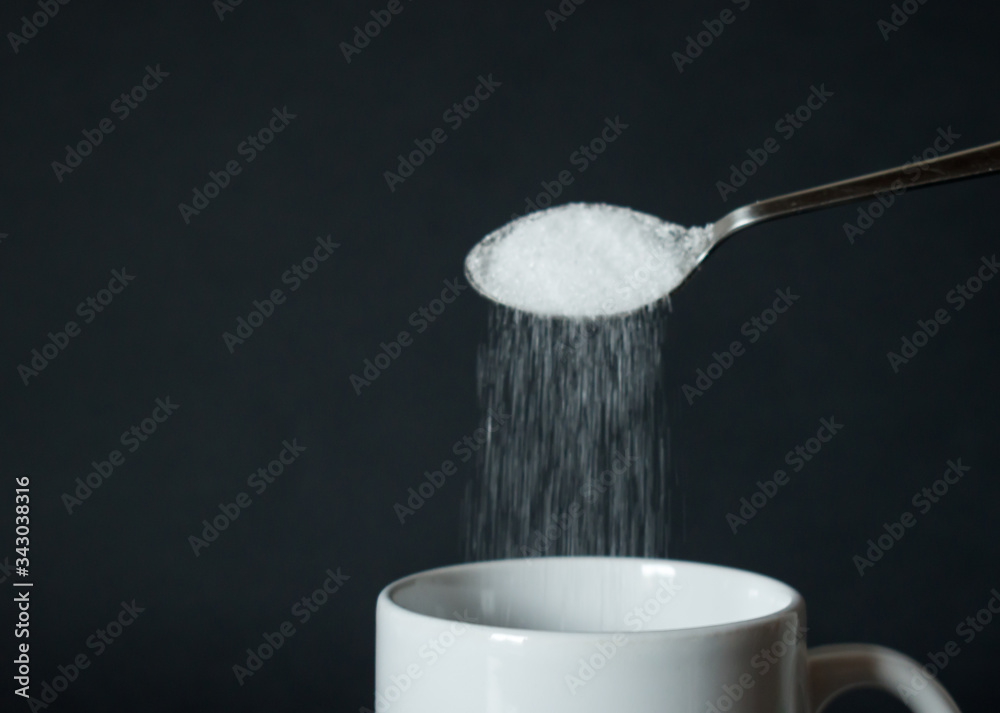 Pouring white sugar from spoon to white coffee cup, mug in front of black background Pouring white sugar from spoon to white coffee cup, mug in front of black background