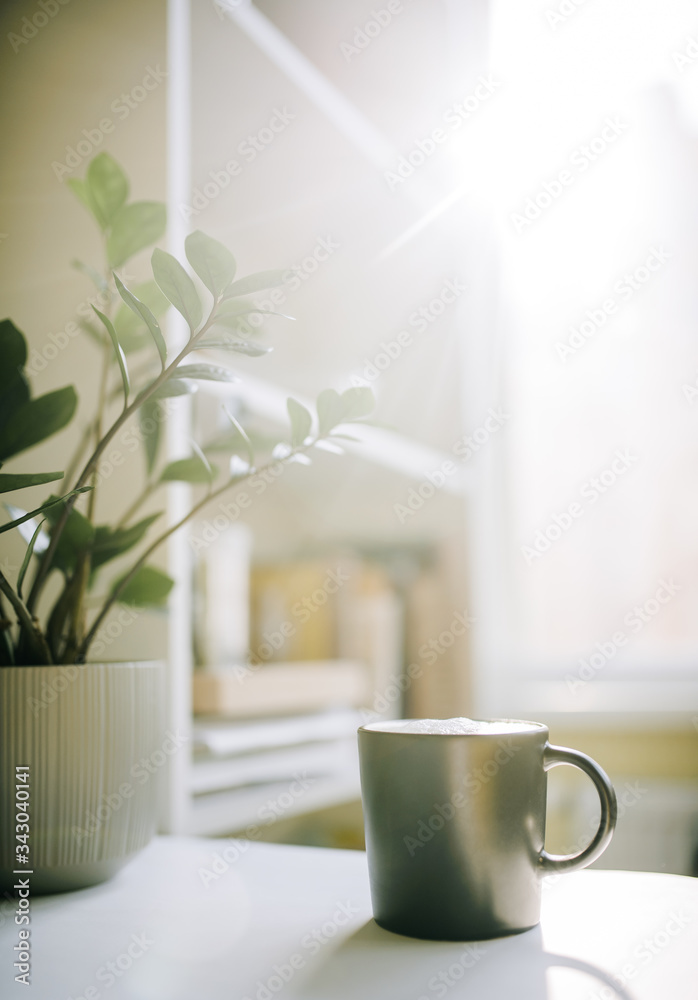A cup of coffee stands on a table near a flowerpot with a plant in the morning window background. Vertical photo A cup of coffee stands on a table near a flowerpot with a plant in the morning window background. Vertical photo