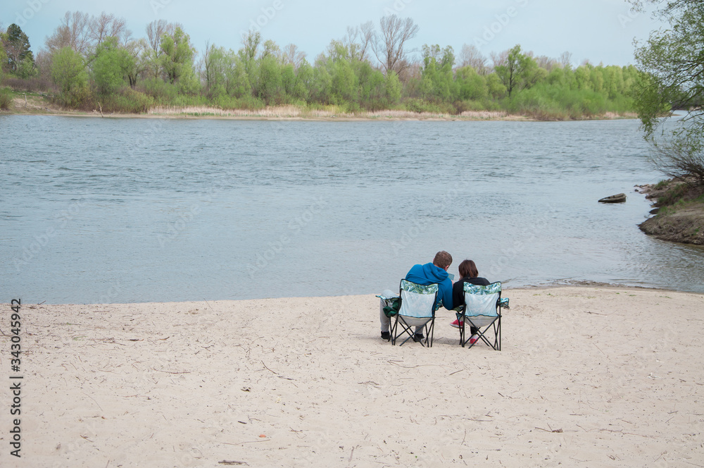 couple man and woman sitting on riverside on chairs in spring and watching film on device and resting on holidays, warm days couple man and woman sitting on riverside on chairs in spring and watching film on device and resting on holidays, warm days