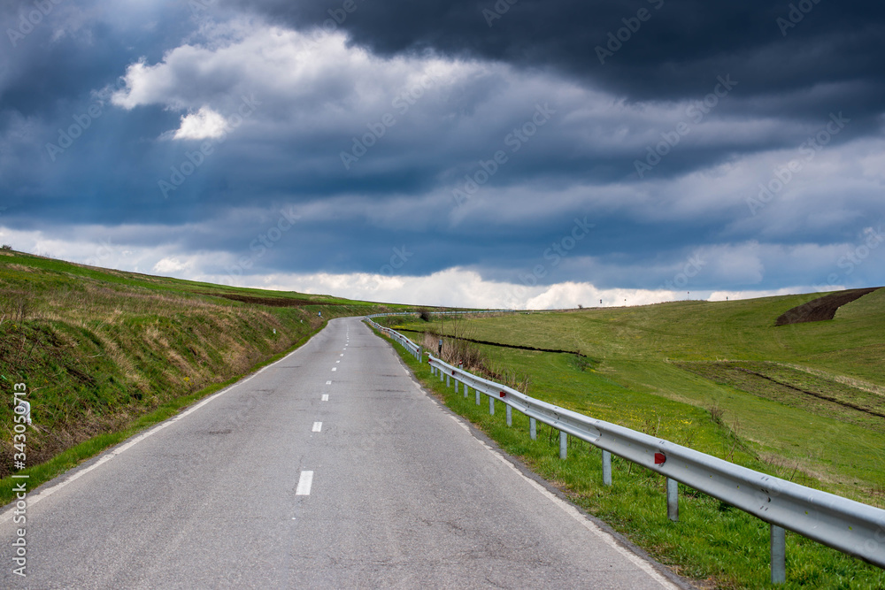 Asphalt road leading to the top of the hill, vibrant green meadow at springtime with dramatic stormclouds.