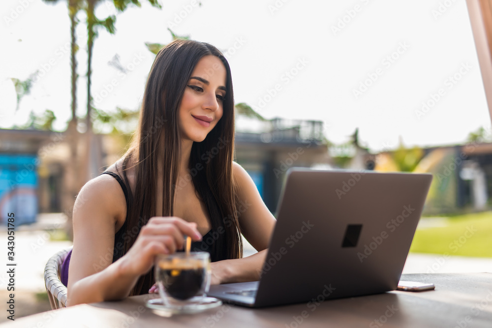 Smiling woman typing on laptop computer and holding coffee cup at the table outdoors Smiling woman typing on laptop computer and holding coffee cup at the table outdoors