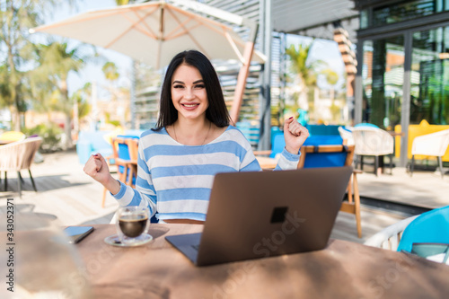 Woman with laptop pumping fists happy laughing online lottery winner. . Girl with long hair sitting outdoors on a coffee shop terrace happy euphoric.