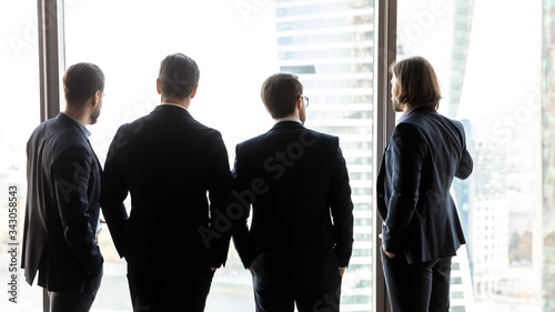 Rear view close up four confident successful businessmen looking through large panoramic window, standing in modern office, enjoying cityscape, thinking about strategy, business vision concept