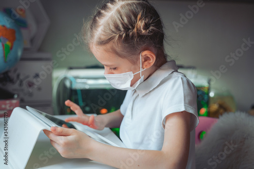 Caucasian preteen girl with medical mask on her face concentrated on her task with tablet. Concept of distance learning in isolation while coronavirus.