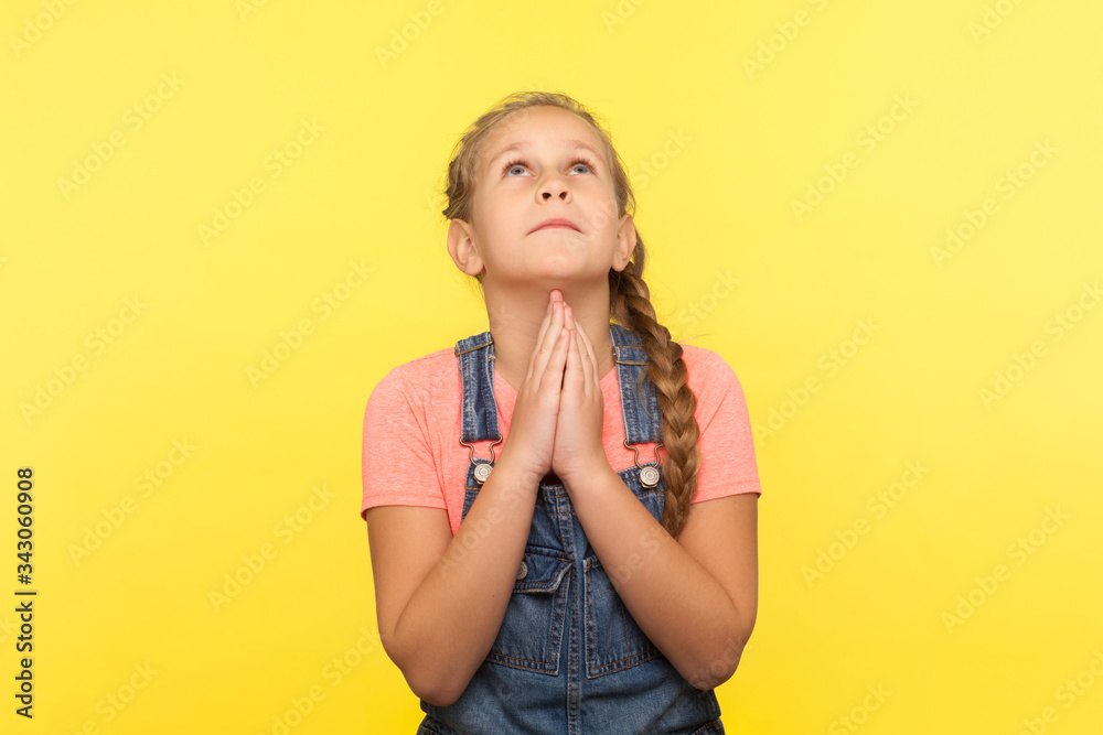 Portrait of imploring little girl in denim overalls looking up praying to god with pleading eyes, asking heartily, making childhood wish and hoping. indoor studio shot isolated on yellow background Portrait of imploring little girl in denim overalls looking up praying to god with pleading eyes, asking heartily, making childhood wish and hoping. indoor studio shot isolated on yellow background