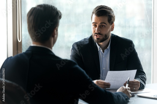 Confident hr manager asking questions to candidate on job interview, sitting at desk, holding cv in hands, business partners discussing project strategy, team leader giving instructions to intern