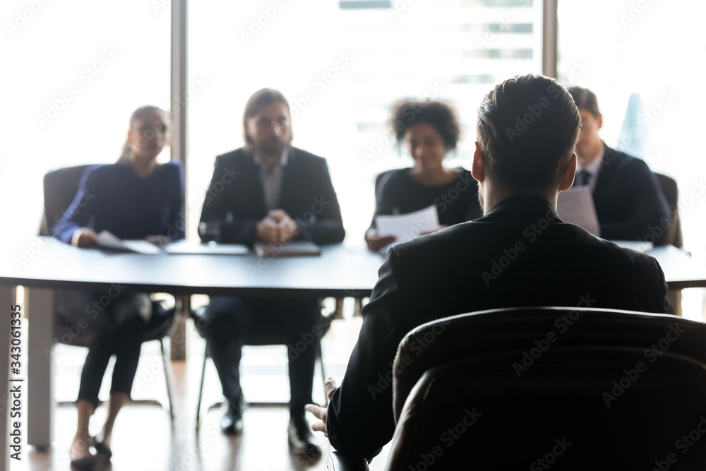 Rear view candidate waiting for hr managers decision, interview result, sitting in front of four diverse recruiters in modern boardroom, male applicant applying for vacancy, employment concept
