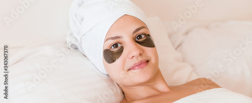 Young woman with black patches under her eyes is lying and relaxing in the bed after having a bath wrapped in towel. Beauty treatment and skincare concept