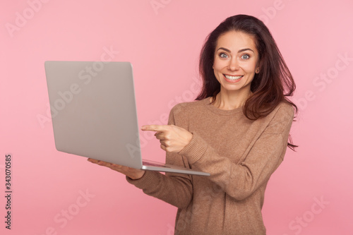 Portrait of amazed woman pointing at laptop and looking at camera with shocked expression, pleasantly surprised by computer software, shopping online. indoor studio shot isolated on pink background