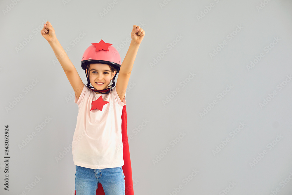 Little baby superhero in a helmet smiles raised his hands while standing against a gray background. Little baby superhero in a helmet smiles raised his hands while standing against a gray background.