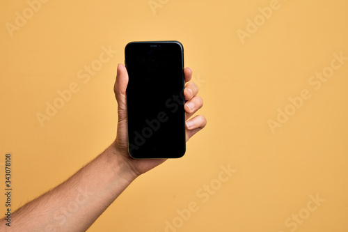 Hand of caucasian young man holding smartphone showing screen over isolated yellow background