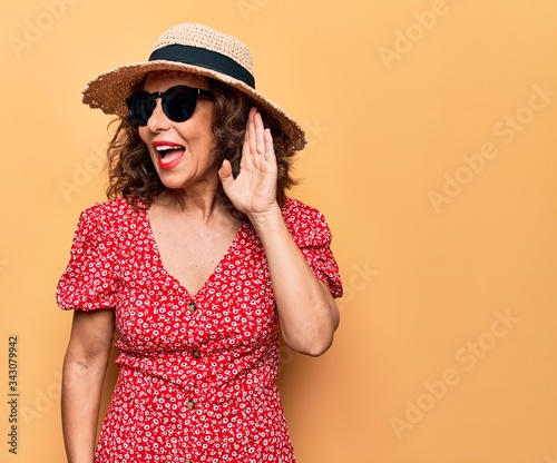 Middle age beautiful woman wearing casual dress and sunglasses over yellow background smiling with hand over ear listening and hearing to rumor or gossip. Deafness concept.