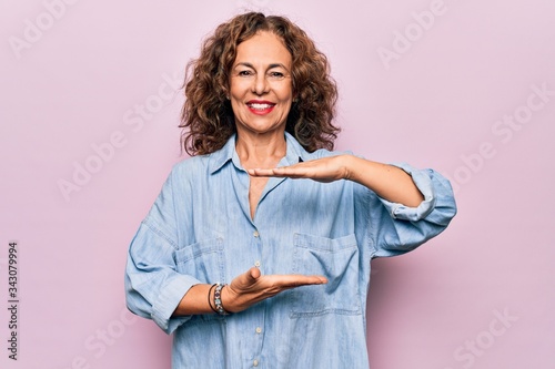 Middle age beautiful woman wearing casual denim shirt standing over pink background gesturing with hands showing big and large size sign, measure symbol. Smiling looking at the camera. Measuring