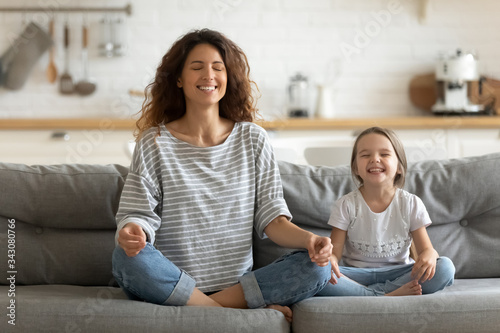 Full length happy young nanny mom sitting on sofa in lotus pose, teaching small daughter yoga breathing exercise. Friendly sincere two female generations family relaxing on couch, healthcare concept.