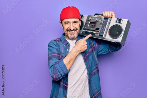 Middle age man wearing cap listening vintage boombox over isolated purple background smiling happy pointing with hand and finger
