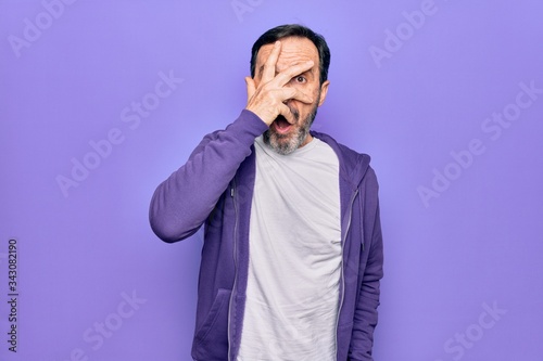 Middle age handsome sporty man wearing casual purple sweatshirt over isolated background peeking in shock covering face and eyes with hand, looking through fingers afraid