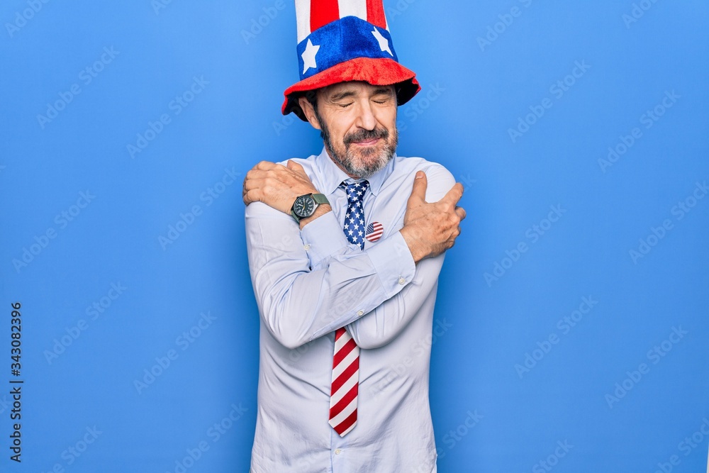 Middle age handsome patriotic man wearing united states hat and tie over blue background hugging oneself happy and positive, smiling confident. Self love and self care Middle age handsome patriotic man wearing united states hat and tie over blue background hugging oneself happy and positive, smiling confident. Self love and self care