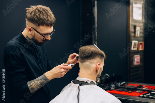 Making haircut look perfect. Young bearded man getting haircut by hairdresser while sitting in chair at barbershop. tattoo