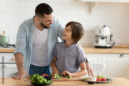 Smiling young father teaching little kid son chopping fresh vegetables for healthy vegetarian dinner, enjoying spending time together in kitchen. Happy child boy learning cooking with dad at home.