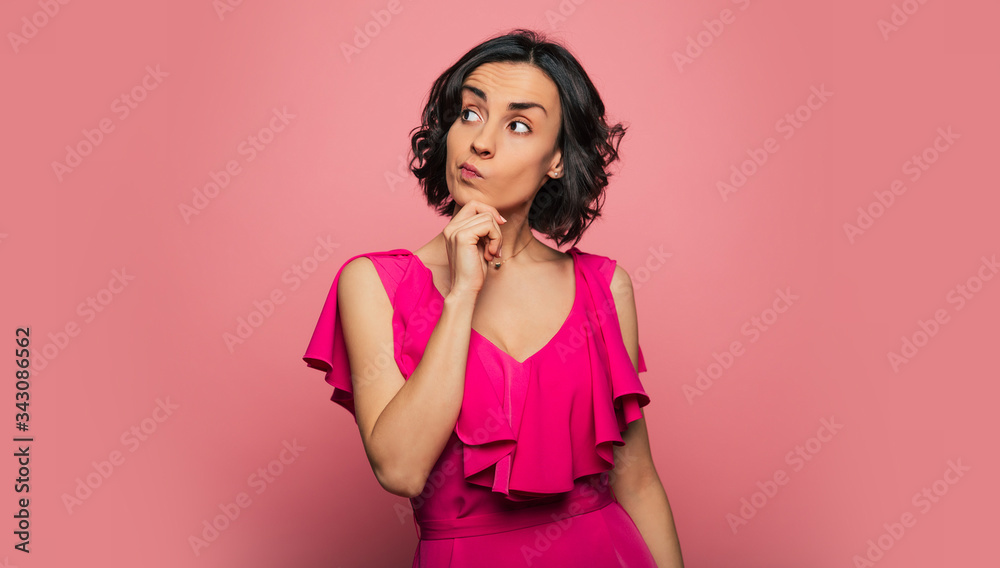 Hard choice. Close-up photo of a gorgeous brunette lady in a pink dress, who is looking to the right and touching her chin with her right hand, thinking over something. Hard choice. Close-up photo of a gorgeous brunette lady in a pink dress, who is looking to the right and touching her chin with her right hand, thinking over something.