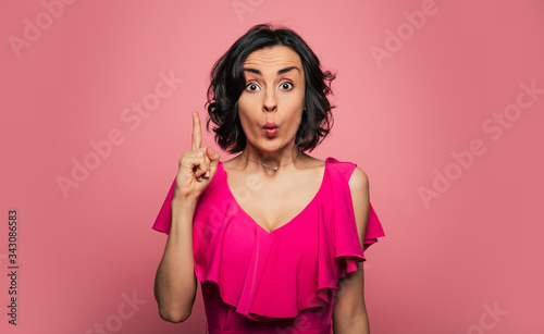 Sheer surprise. Close-up photo of a surprised young woman in a pink dress  who is pointing upwards with her right index finger.