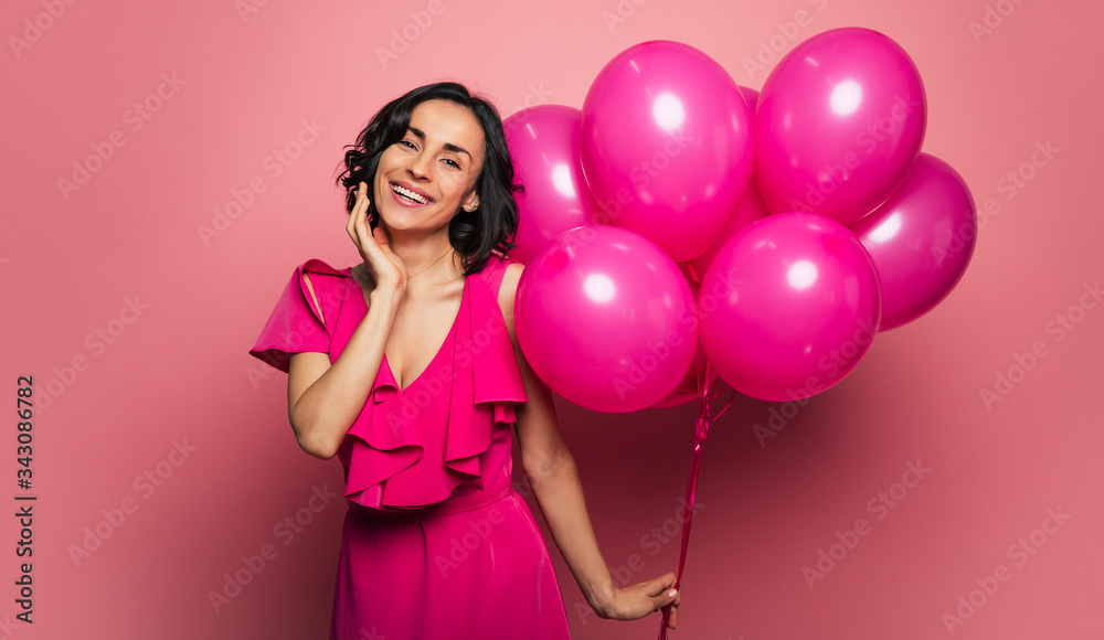 Happy birthday to me! Young adult woman in a festive dress is holding a bunch of balloons in her left hand and smiling broadly, while celebrating her birthday.