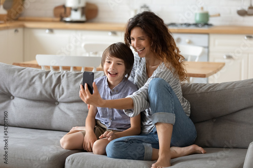 Joyful young woman holding mobile phone, taking selfie shot with small cute kid boy. Smiling female blogger recording funny video vlog with adorable child son, spending time together at home.