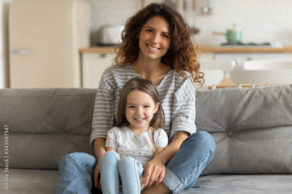 Portrait of mother relaxing on couch with little preschool child daughter. Happy young woman embracing small kid girl, looking at camera. Smiling two female generations posing for family photo. Portrait of mother relaxing on couch with little preschool child daughter. Happy young woman embracing small kid girl, looking at camera. Smiling two female generations posing for family photo.