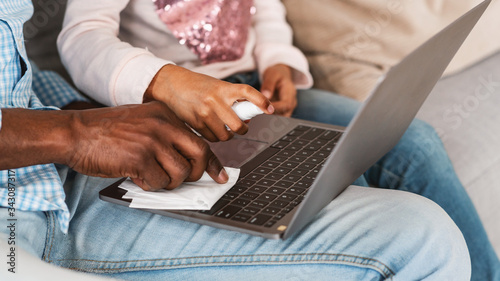 African American man with grandchild using sanitizer to clean laptop at home, closeup. Panorama