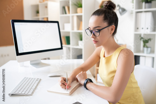 Busy young woman with hair bun sitting at desk with computer and writing down in diary while planning tasks at work