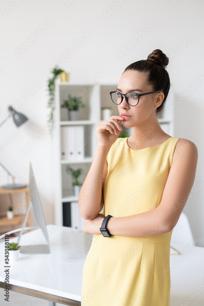 Pensive enterprising young businesswoman in yellow dress and eyeglasses standing with hand near lips in modern office