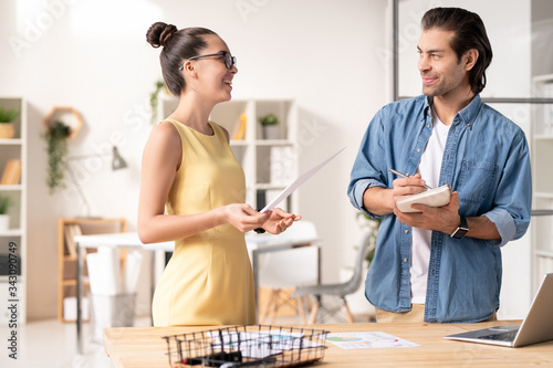 Positive young businesswoman in yellow dress standing by desk with laptop and analyzing sales report with colleague