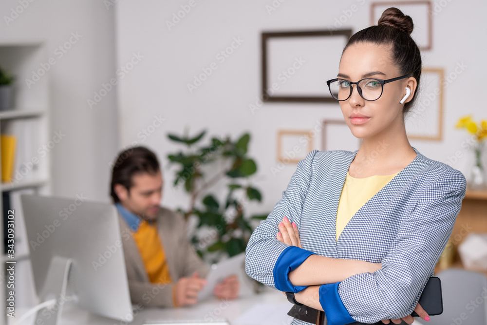 Portrait of stylish young businesswoman in wireless ear buds standing with crossed arms in modern office, man working in background Portrait of stylish young businesswoman in wireless ear buds standing with crossed arms in modern office, man working in background