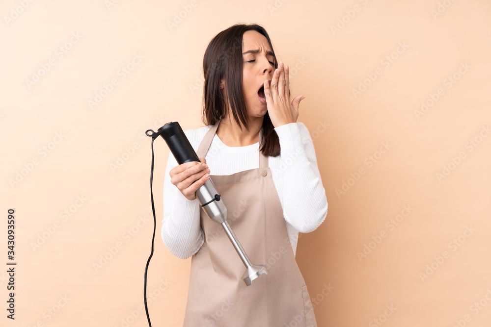 Young brunette girl using hand blender over isolated background yawning and covering wide open mouth with hand