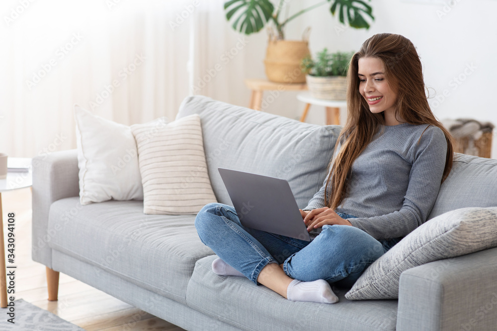 Smiling young lady sitting with laptop on couch at home - obrazy, fototapety, plakaty 