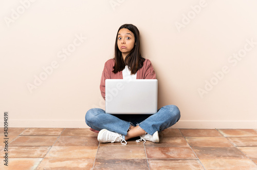 Young mixed race woman with a laptop sitting on the floor making doubts gesture while lifting the shoulders © luismolinero