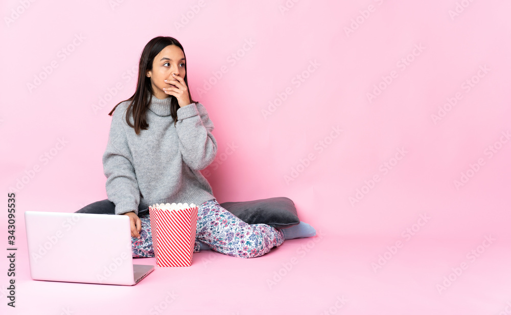Young mixed race woman eating popcorn while watching a movie on the laptop doing surprise gesture while looking to the side Young mixed race woman eating popcorn while watching a movie on the laptop doing surprise gesture while looking to the side