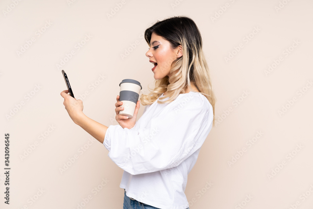 Teenager girl isolated on beige background holding coffee to take away and a mobile Teenager girl isolated on beige background holding coffee to take away and a mobile