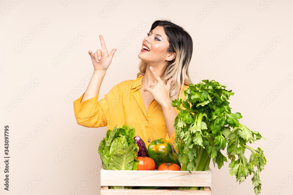 Teenager farmer girl with freshly picked vegetables in a box isolated on beige background pointing with the index finger a great idea Teenager farmer girl with freshly picked vegetables in a box isolated on beige background pointing with the index finger a great idea