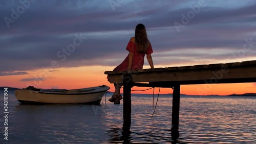 Young slender girl with long blonde hair, in red dress with bare shoulders, sits with her back on a wooden pier or bridge and looks at a beautiful sunset and a wooden boat on water. Relax, loneliness.