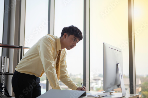 Young businessmen are deliberately writing down important information displayed on a computer screen in a notebook to present the executive board at the next meeting.