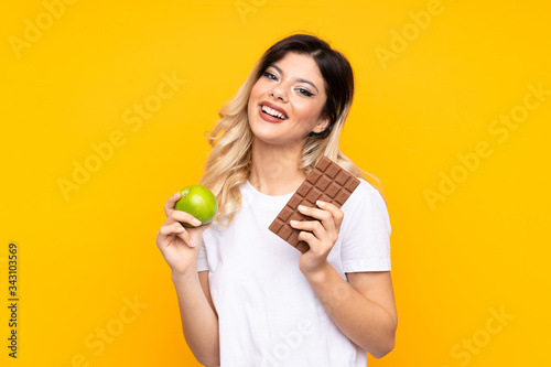 Teenager girl isolated on yellow background having doubts while taking a chocolate tablet in one hand and an apple in the other