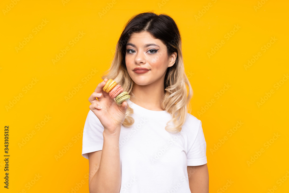 Teenager girl isolated on yellow background holding colorful French macarons and with sad expression Teenager girl isolated on yellow background holding colorful French macarons and with sad expression