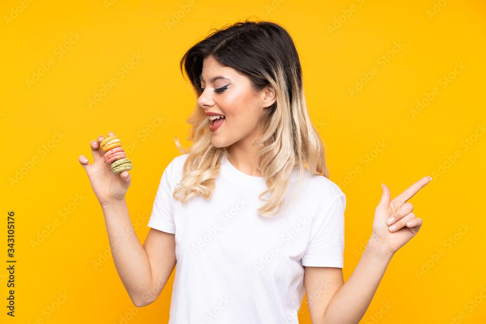 Teenager girl isolated on yellow background holding colorful French macarons and pointing side Teenager girl isolated on yellow background holding colorful French macarons and pointing side