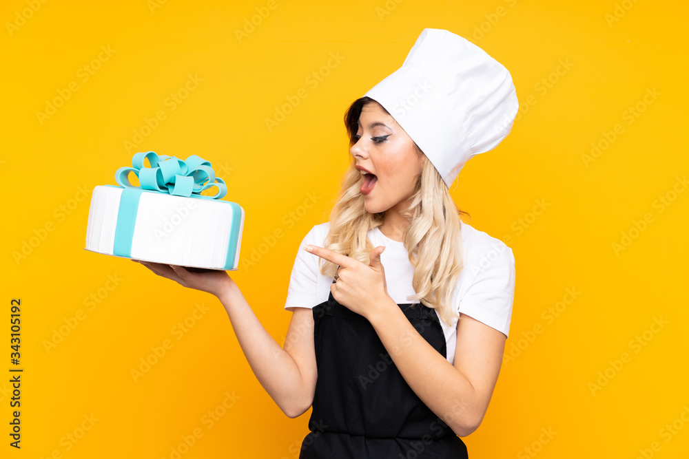 Teenager girl pastry holding a big cake isolated on yellow background surprised and pointing finger to the side Teenager girl pastry holding a big cake isolated on yellow background surprised and pointing finger to the side