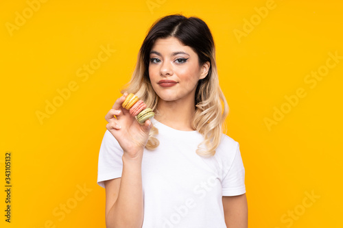 Teenager girl isolated on yellow background holding colorful French macarons and with sad expression